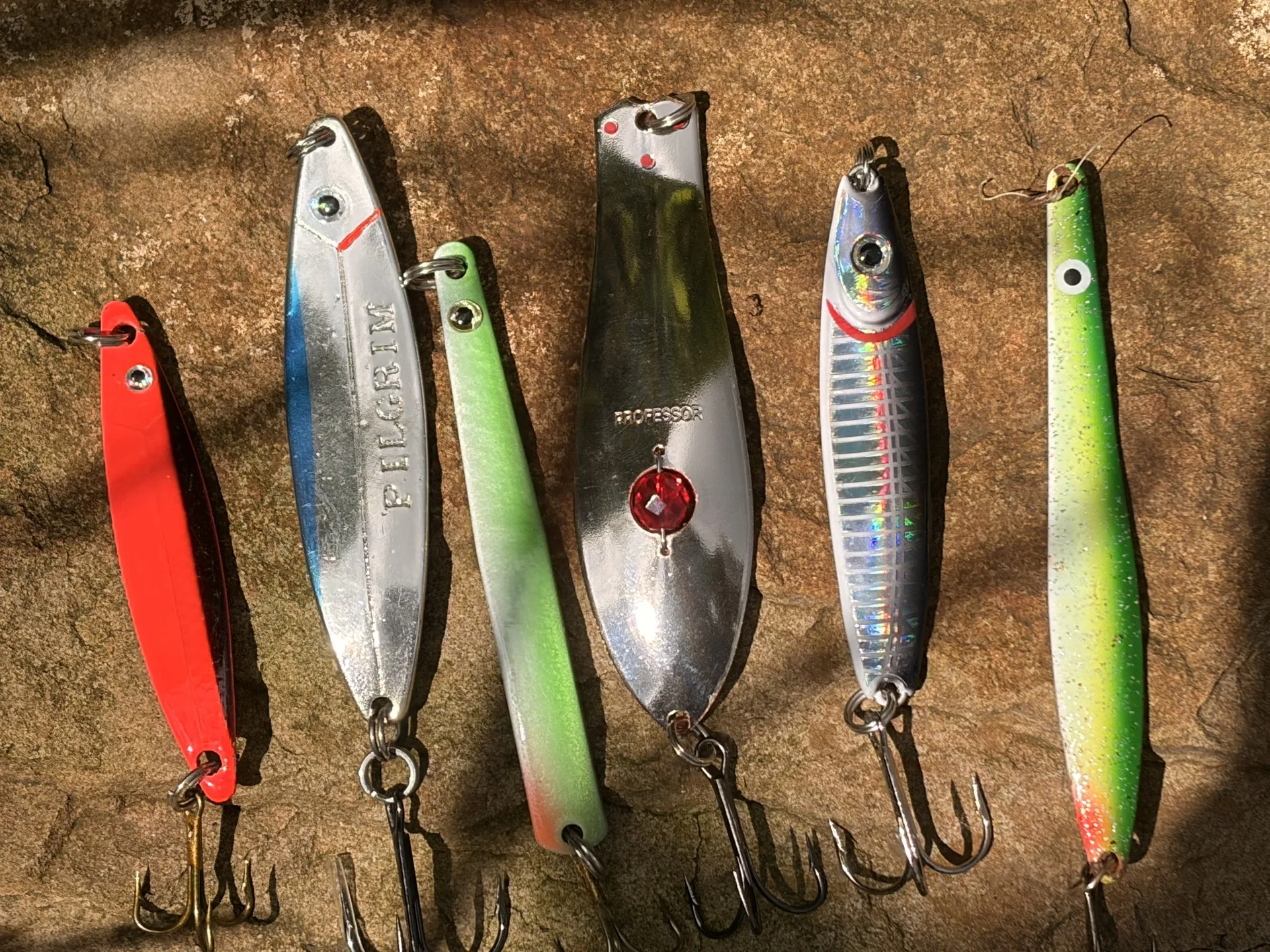 Sechs Meerforellenblinker nebeneinander auf einem Stein: Rot-Schwarz, Silber-Blau, Grün-Weiß, blankes Silber, Blau-Silber und Grün-Gelb
